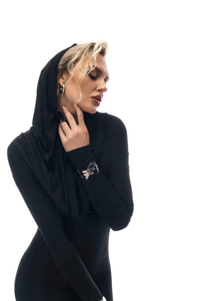 Модель у чорному платті KYLIE MAXI DRESS чорного кольору від бренду Her Name стоїть торкаючись правою рукою обличчя
