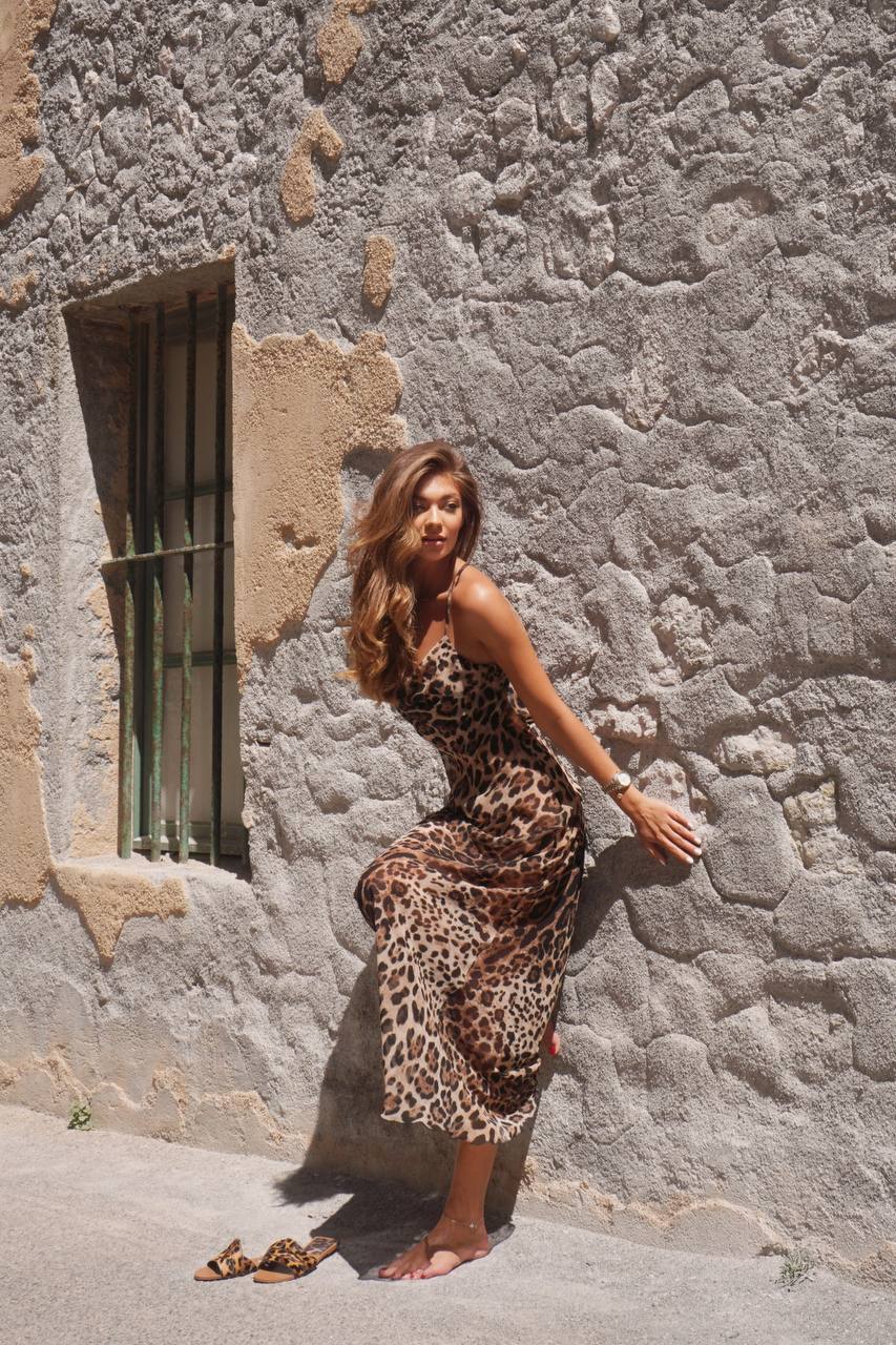 Модель у платті леопардового кольору LEO DRESS від українського бренду HER NAME опирається на стіну камяного будинку.