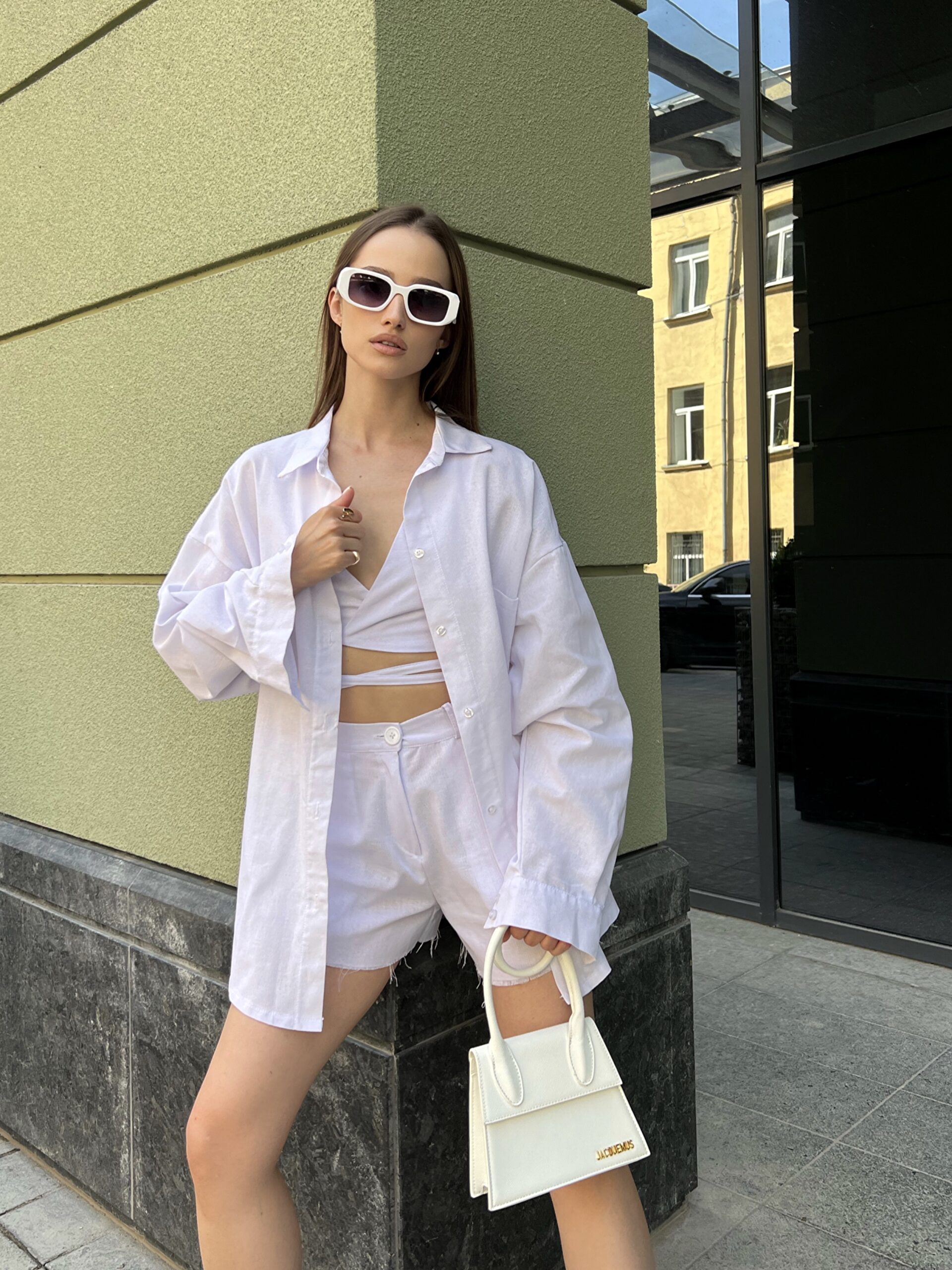 Модель у білій льняній універсальній сорочці та топі бренду Her Name лежить на фоні будівлі стоїть боком в білих окулярах з білою сумкою