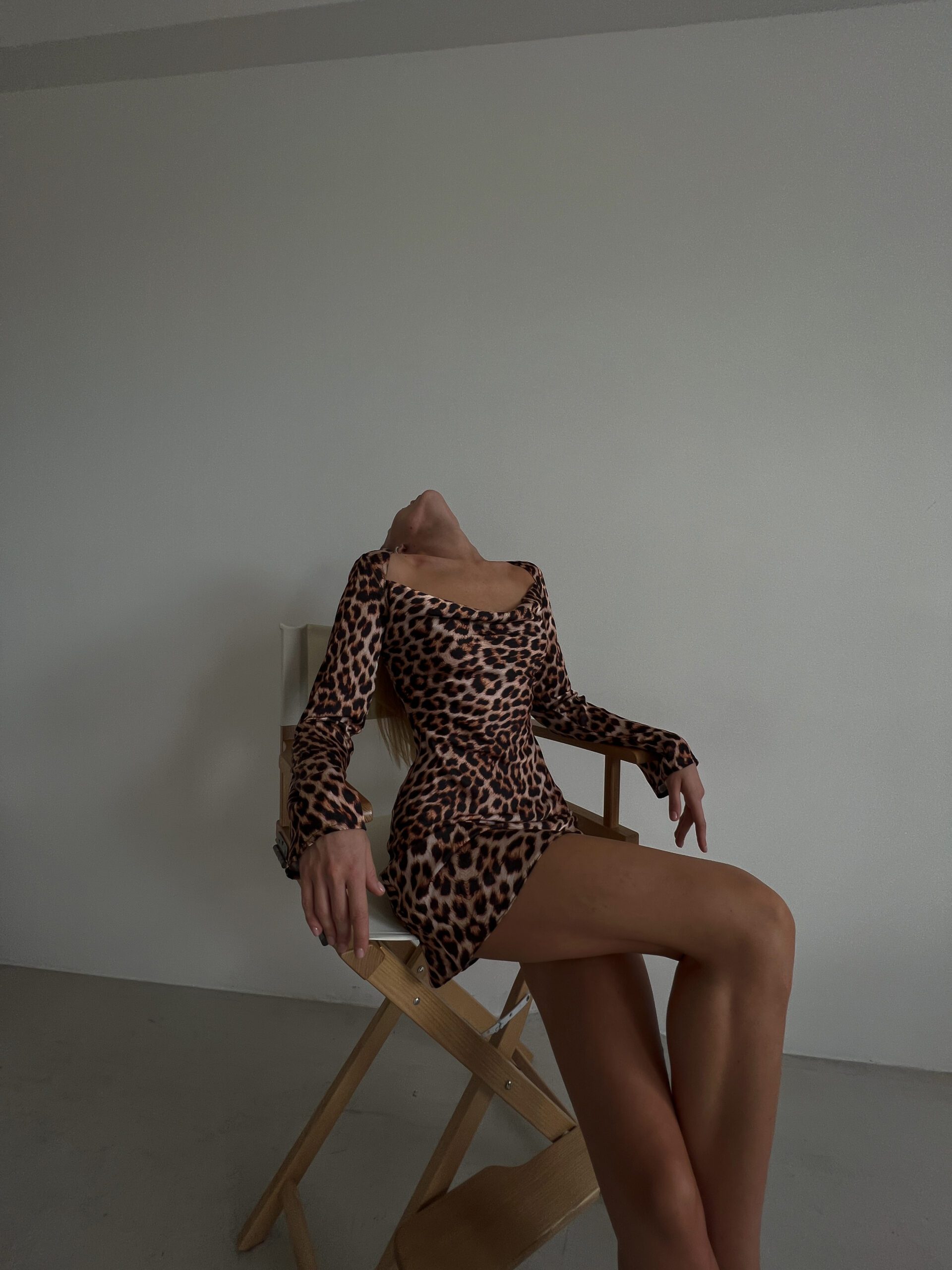 Дівчина у леопардовому платті MINI LEO DRESS бренду Her Name сидить на дерев'яному стільці запрокинувши голову назад