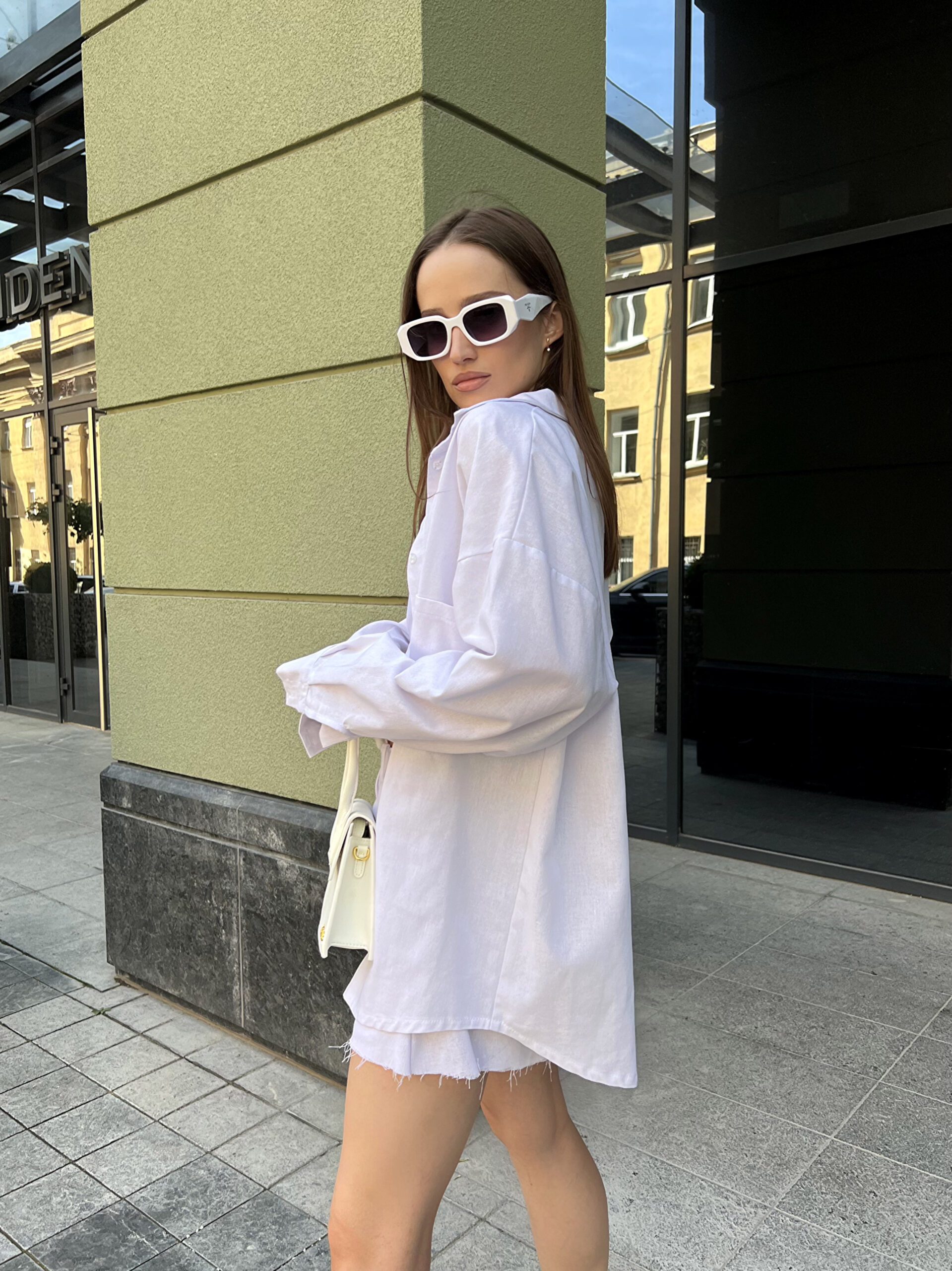 Модель у білій льняній універсальній сорочці бренду Her Name лежить на фоні будівлі стоїть боком в білих окулярах