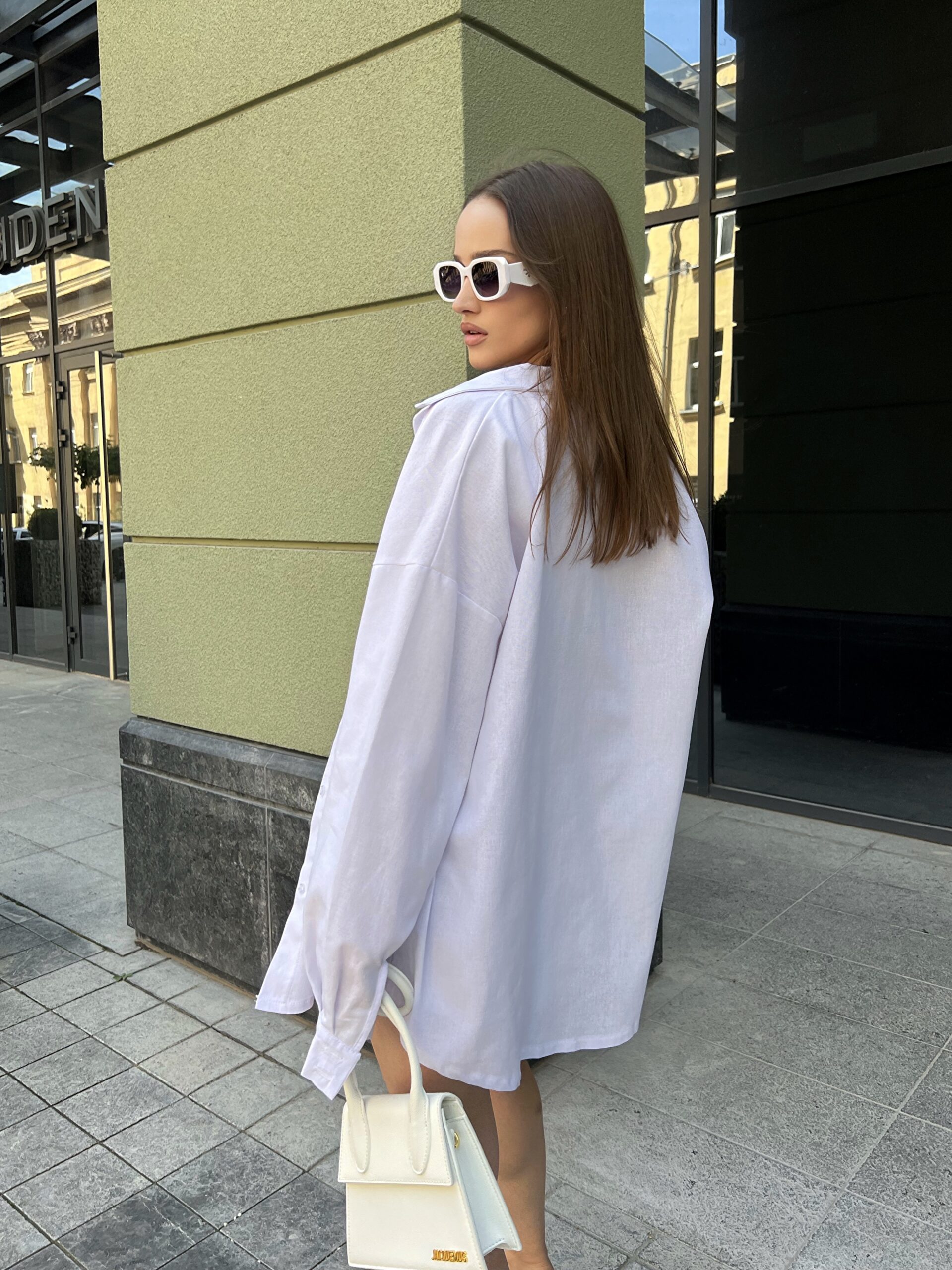 Модель у білій льняній універсальній сорочці та топі бренду Her Name лежить на фоні будівлі стоїть боком в білих окулярах з білою сумкою та шортах стоїть спиною