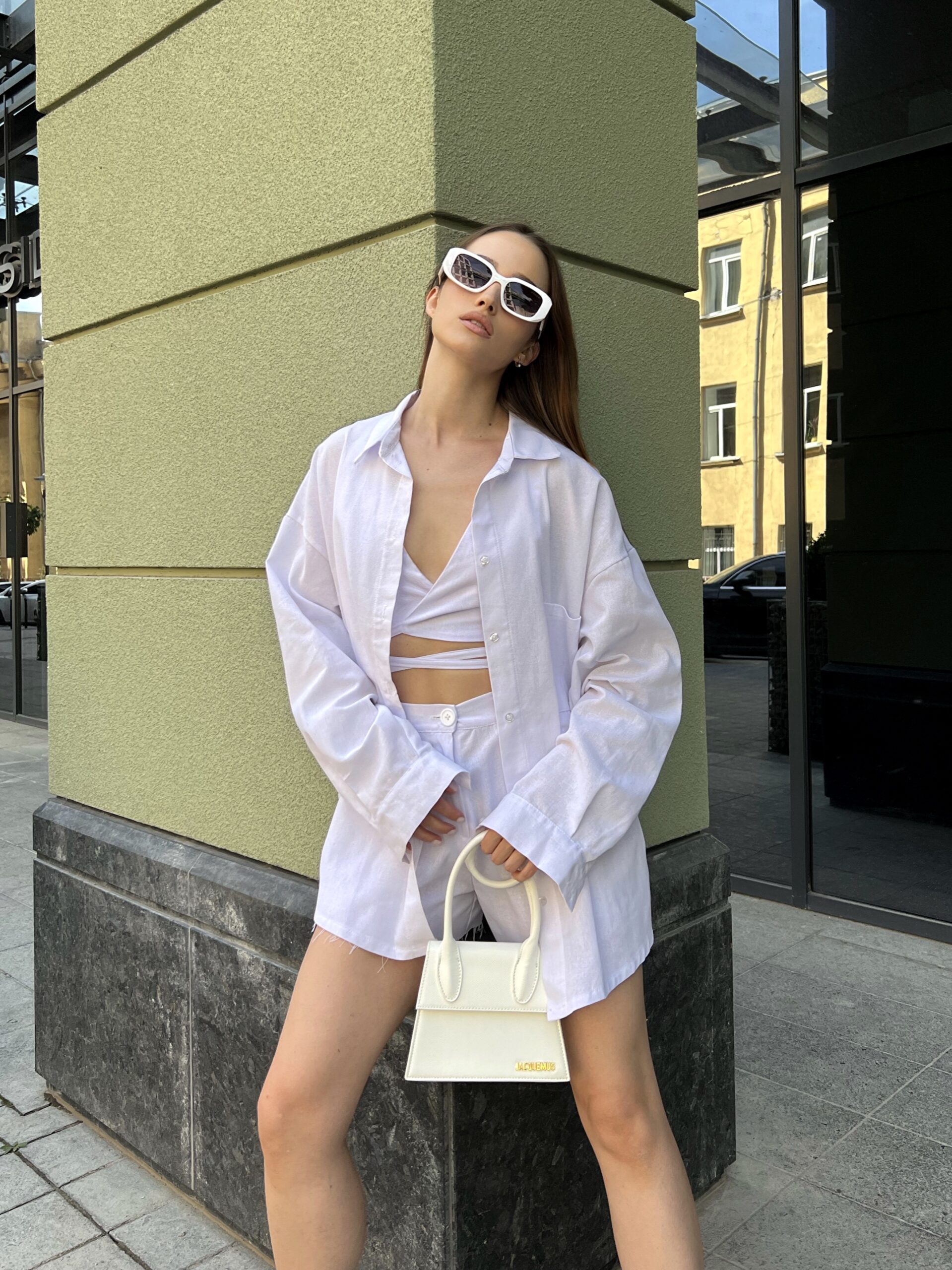 Модель у білій льняній універсальній сорочці бренду Her Name лежить на фоні будівлі стоїть боком в білих окулярах з ілою сумкою
