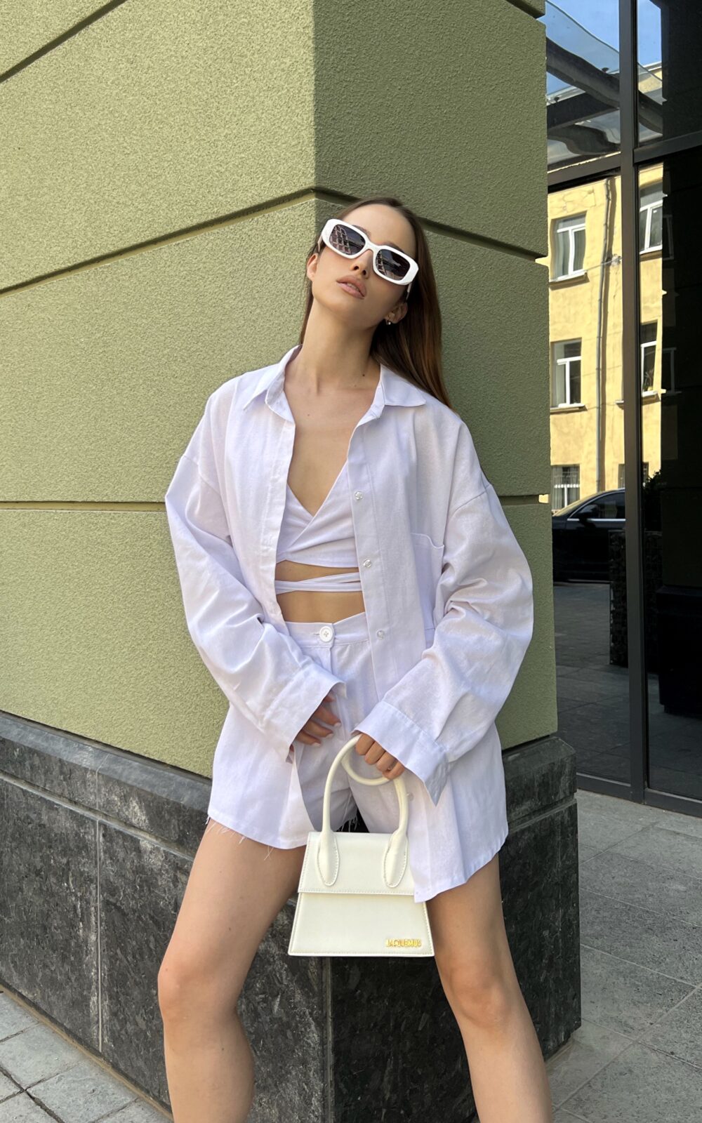 Модель у білій льняній універсальній сорочці бренду Her Name лежить на фоні будівлі стоїть боком в білих окулярах з ілою сумкою