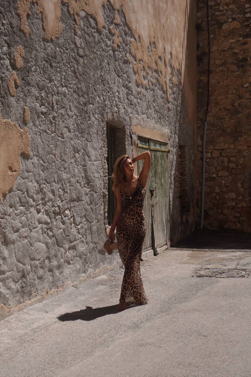 Модель у платті леопардового кольору LEO DRESS від українського бренду HER NAME тримає в руках взуття та прикриває рукою сонце.