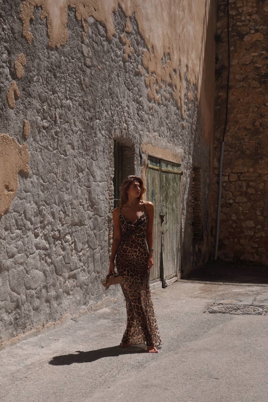 Модель у платті леопардового кольору LEO DRESS від українського бренду HER NAME тримає в руках взуття та дивиться в сторону.