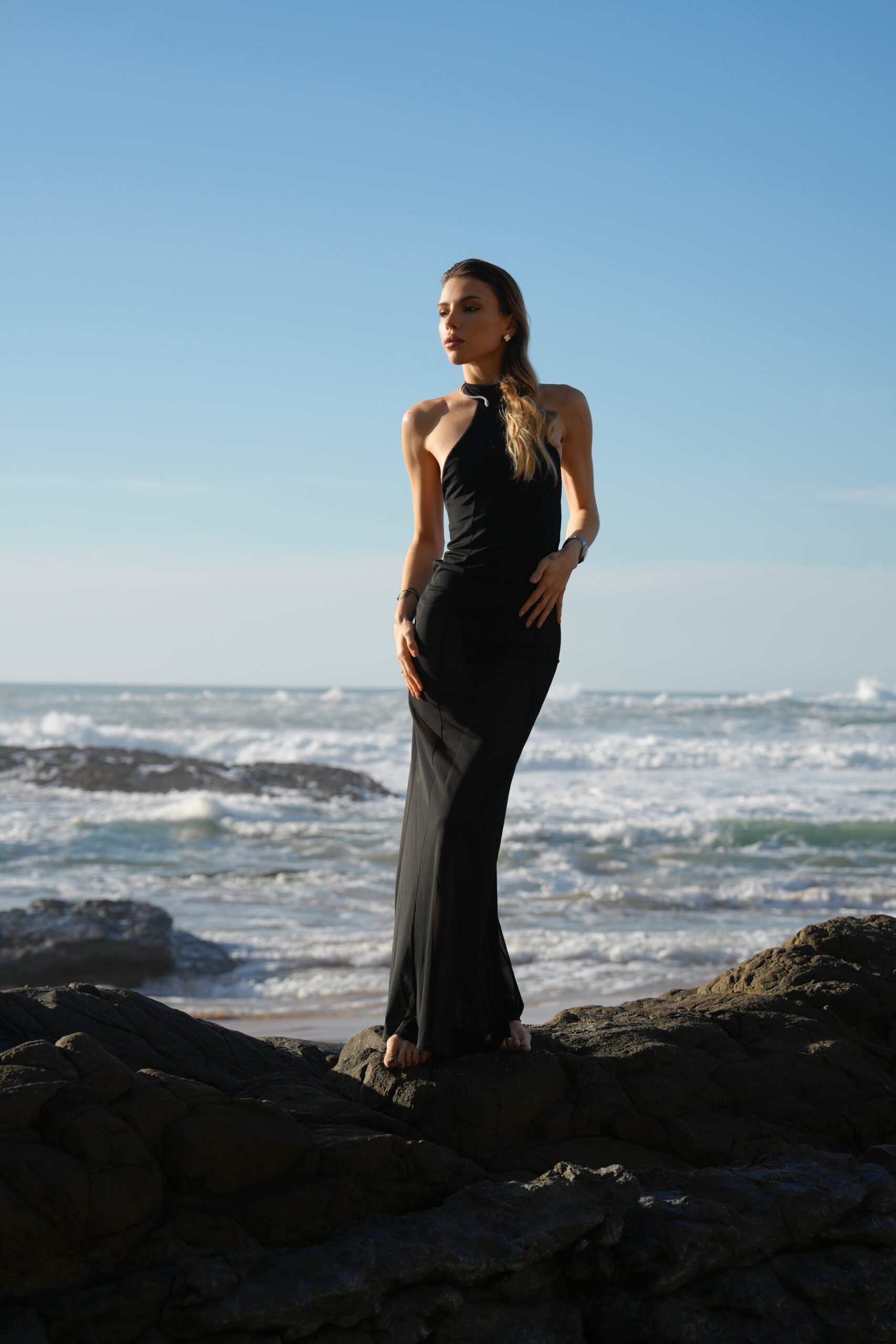 Модель у ANGELINA DRESS (напівпрозорому) платті чорного кольору від бренду Her Name стоїть на камінні на фоні чоного моря