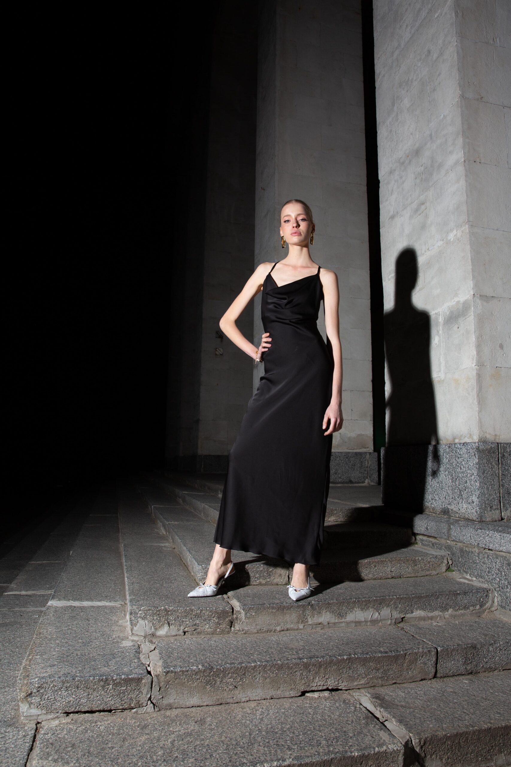 Дівчина у платті чорного кольору SLIP DRESS “LANA” від бренду Her Name стоїть поклавши праву руку на талію