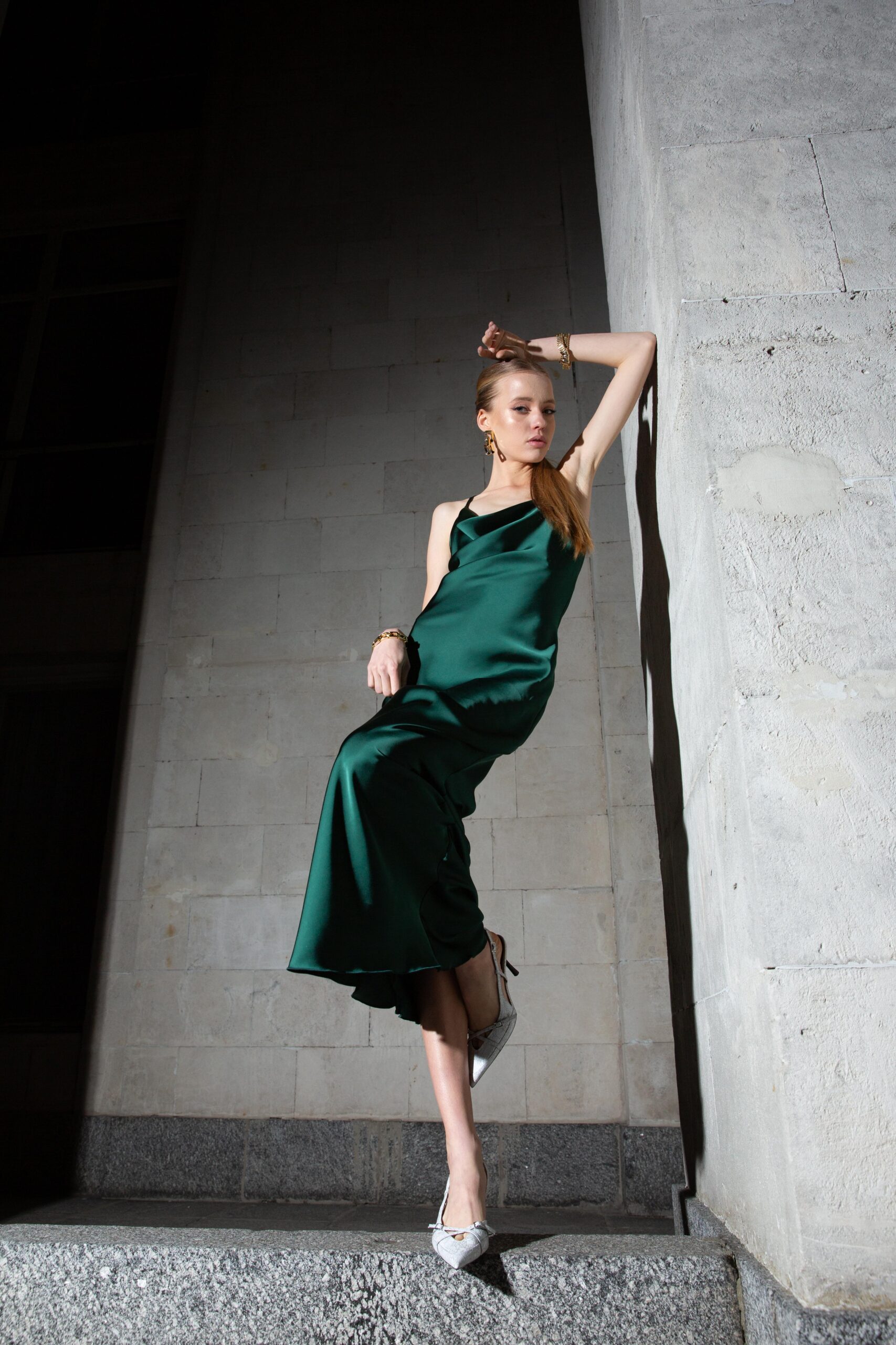 Дівчина у платті смарагдового кольору SLIP DRESS “LANA” від бренду Her Name стоїть спераючись на сіру стіну зігнотую лівою рукою