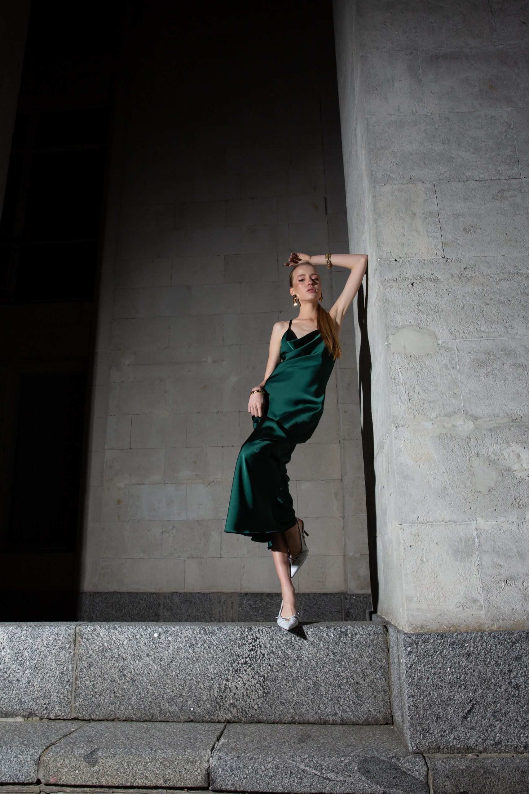 Дівчина у платті смарагдового кольору SLIP DRESS “LANA” від бренду Her Name стоїть спераючись на сіру стіну зігнотую лівою рукою та дивиться прямо