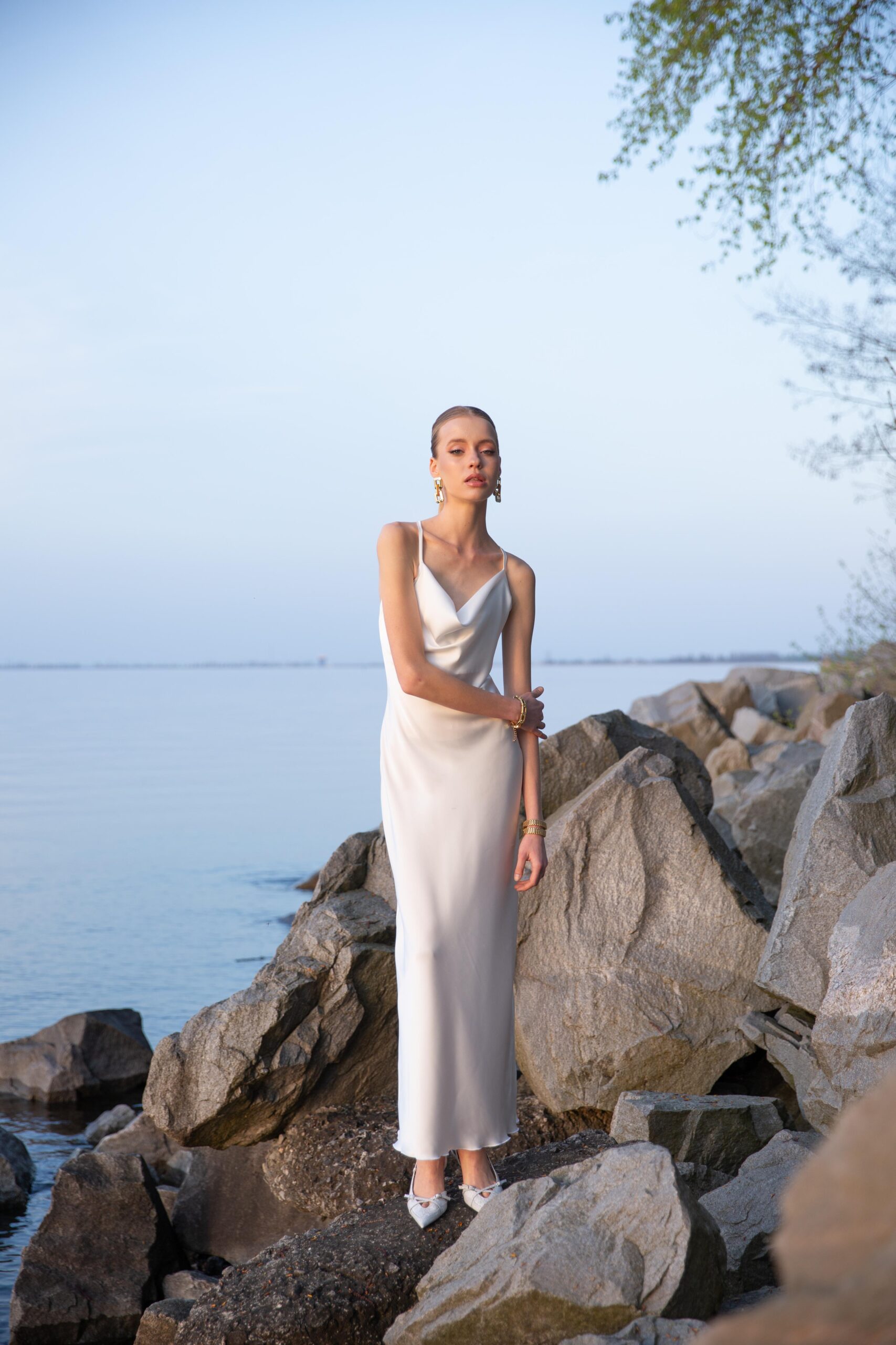 Дівчина у платті білого кольору SLIP DRESS “LANA” від бренду Her Name стоїть на камінні на фоні Чорного моря поклавши праву руку на ліву