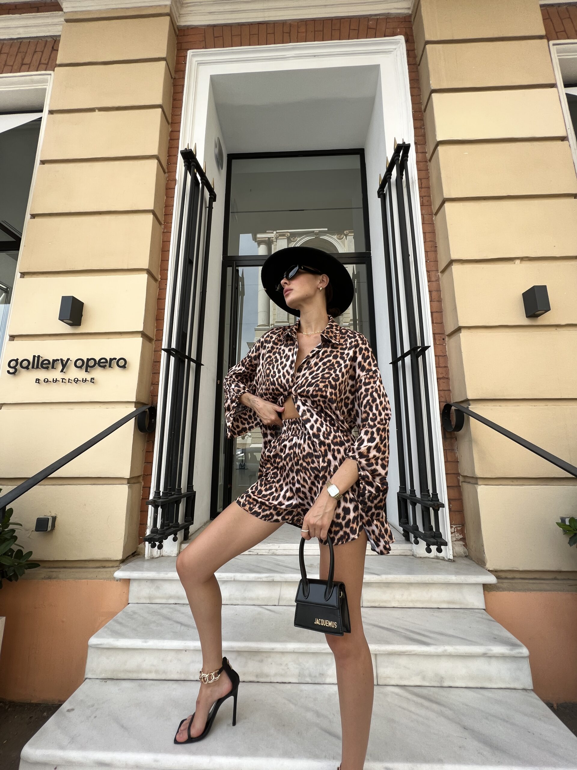 Дівчина у комлекті LEO SUIT лопардового кольору від бренду Her Name на чорних каблуках на мраморних сходах виставивши праву ногу вперед