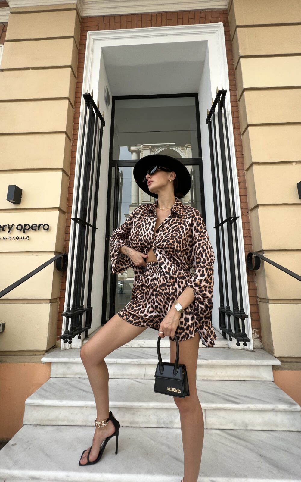 Дівчина у комлекті LEO SUIT лопардового кольору від бренду Her Name на чорних каблуках на мраморних сходах виставивши праву ногу вперед