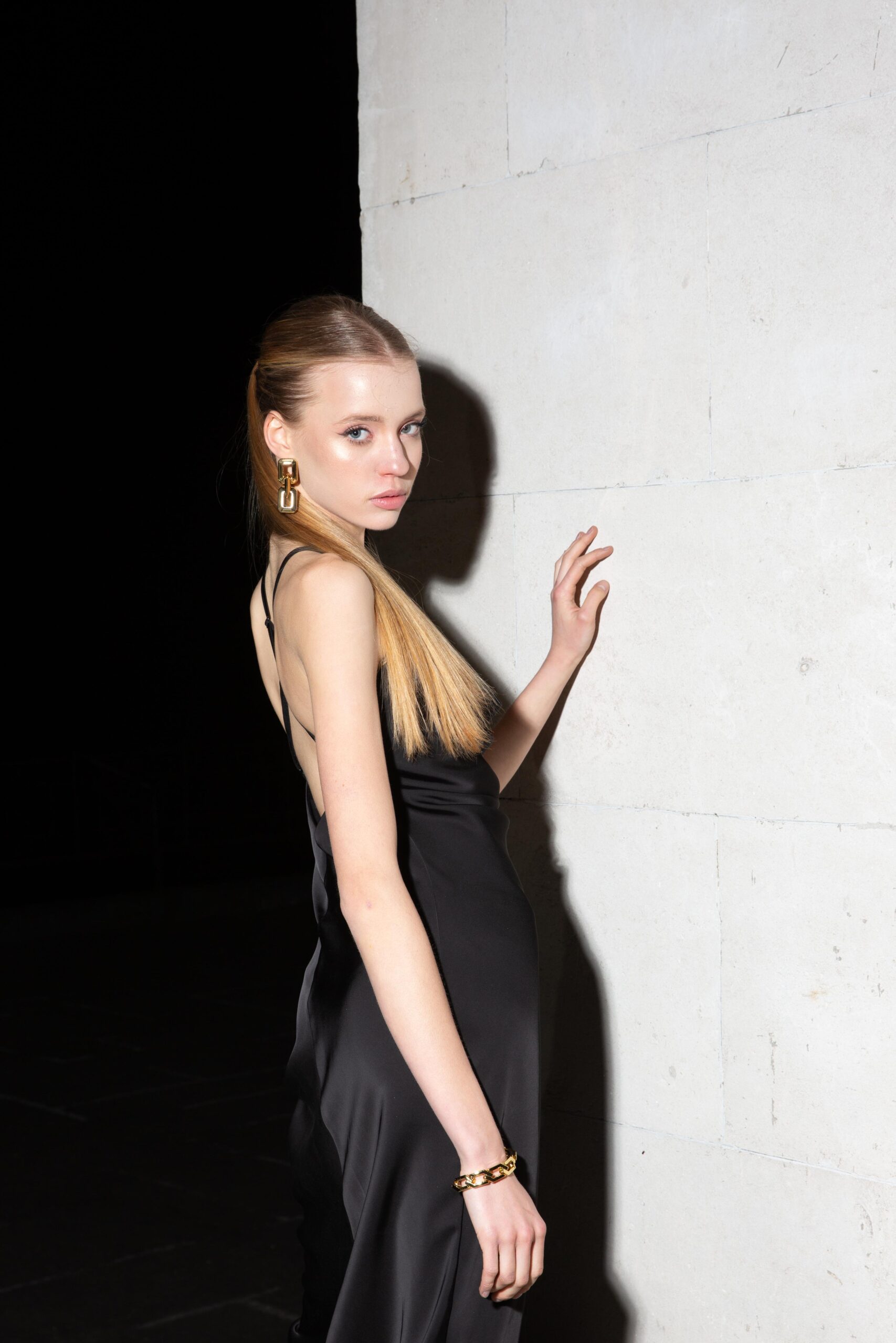 Дівчина у платті чорного кольору SLIP DRESS “LANA” від бренду Her Name стоїть боком біля білої стіни поклавши ліву руку на стіну