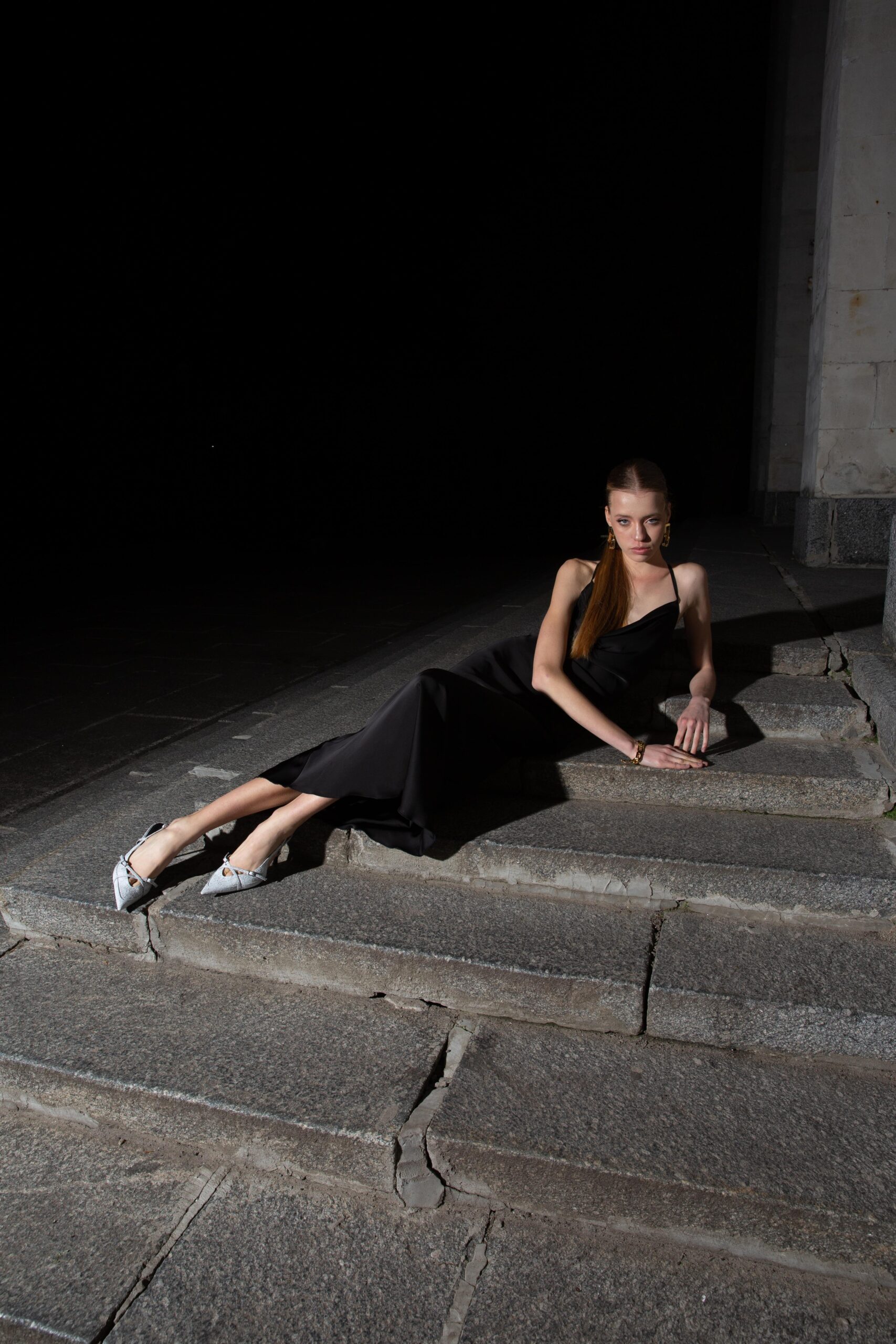 Дівчина у платті чорного кольору SLIP DRESS “LANA” від бренду Her Name лежить на схлдах