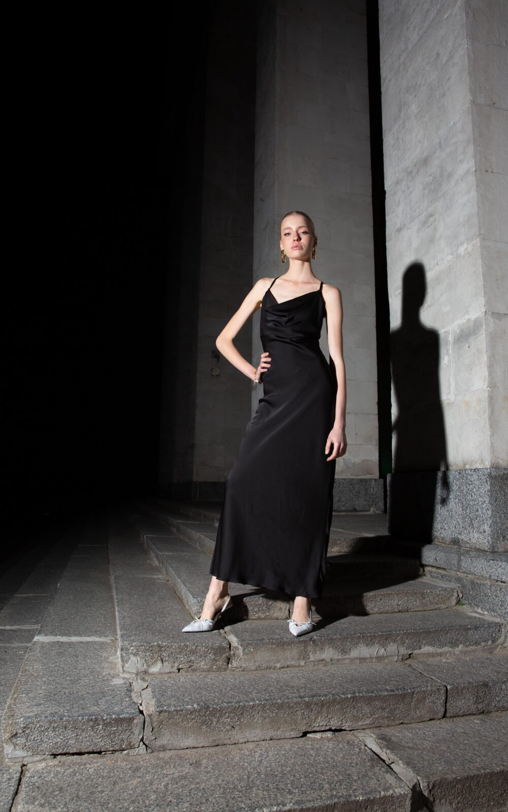 Дівчина у платті чорного кольору SLIP DRESS “LANA” від бренду Her Name стоїть поклавши праву руку на талію