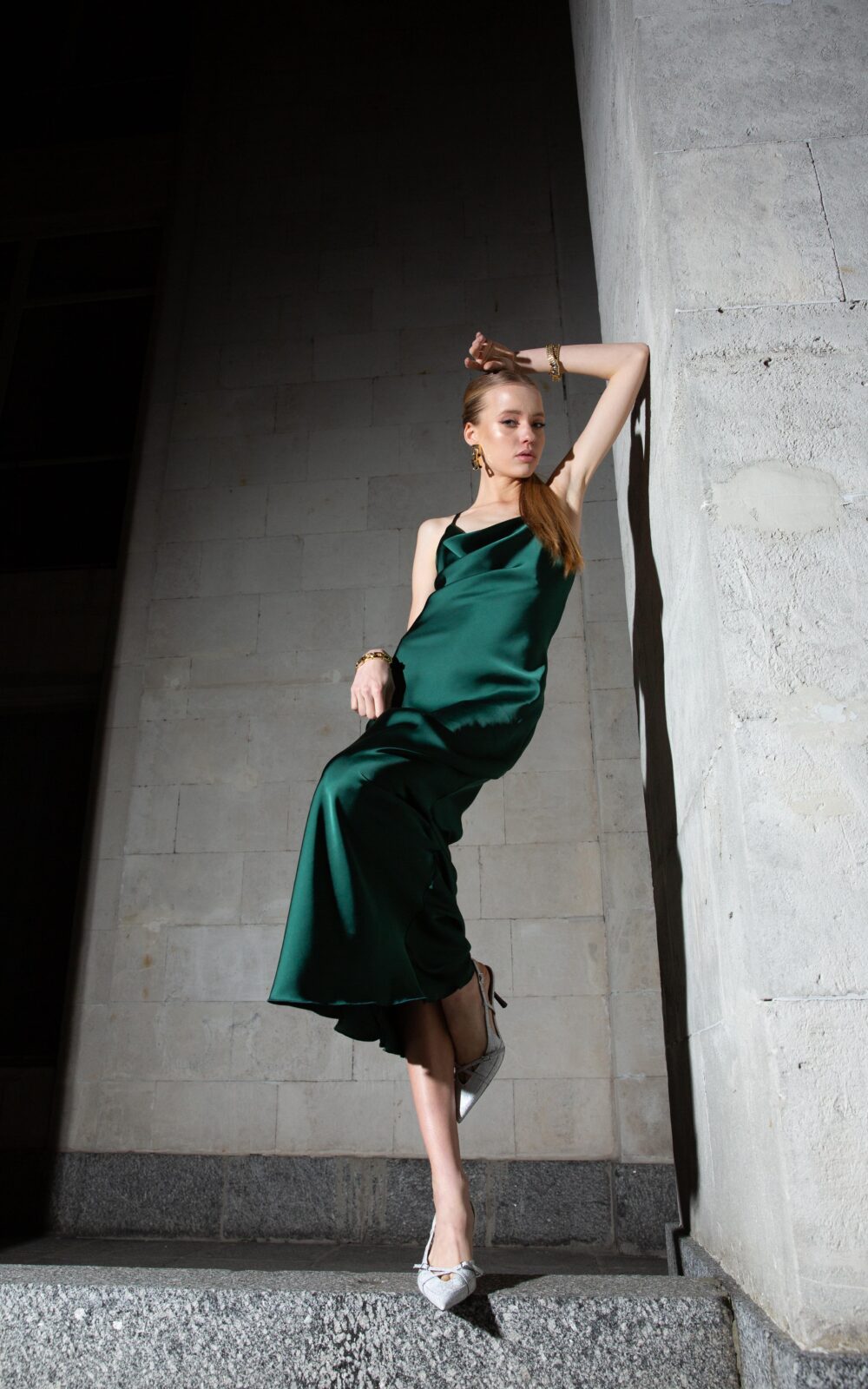 Дівчина у платті смарагдового кольору SLIP DRESS “LANA” від бренду Her Name стоїть спераючись на сіру стіну зігнотую лівою рукою