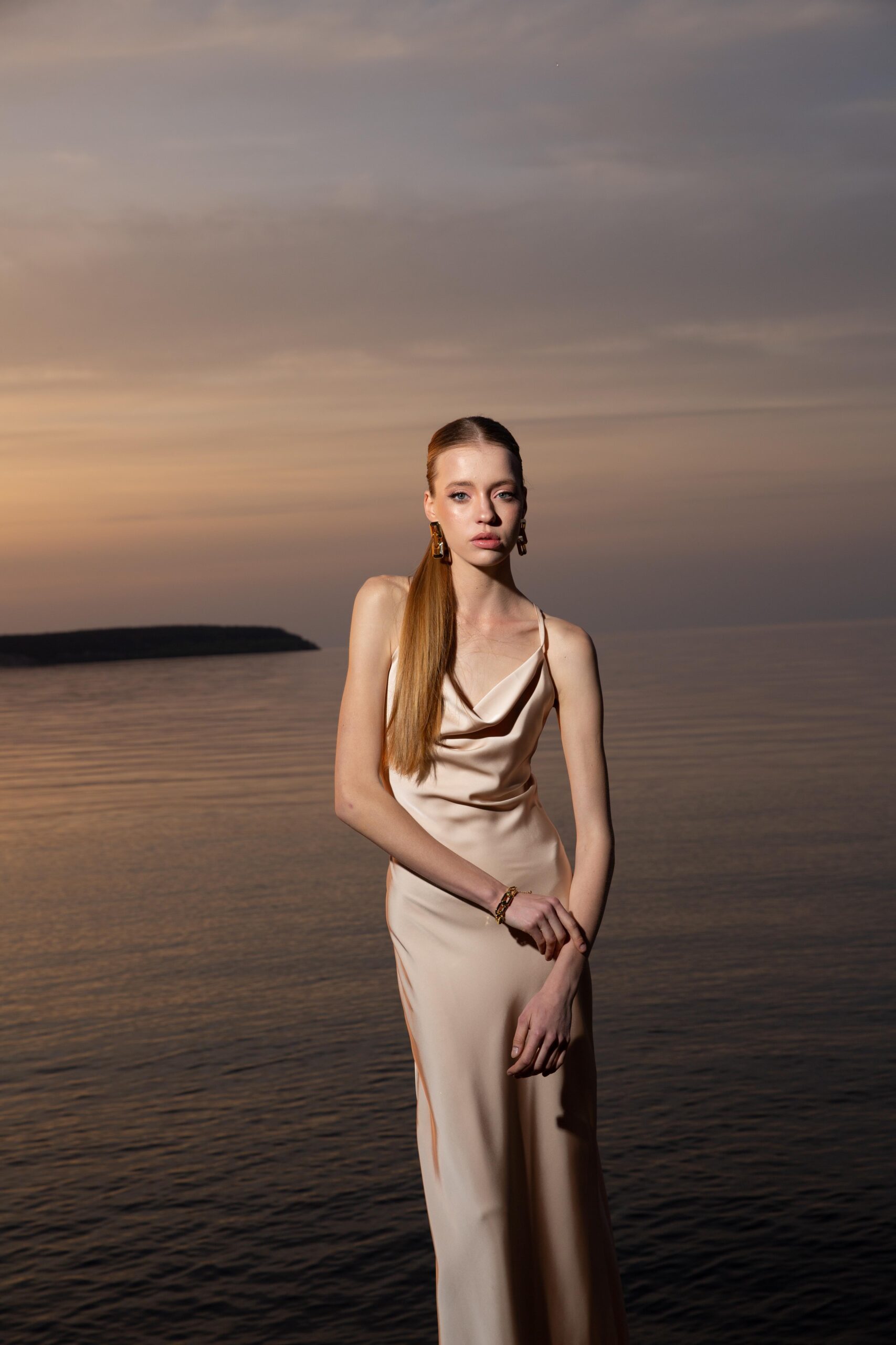 Дівчина у платті шампань кольору SLIP DRESS “LANA” від бренду Her Name стоїть на фоні Чорного моря поклавши праву руку на ліву