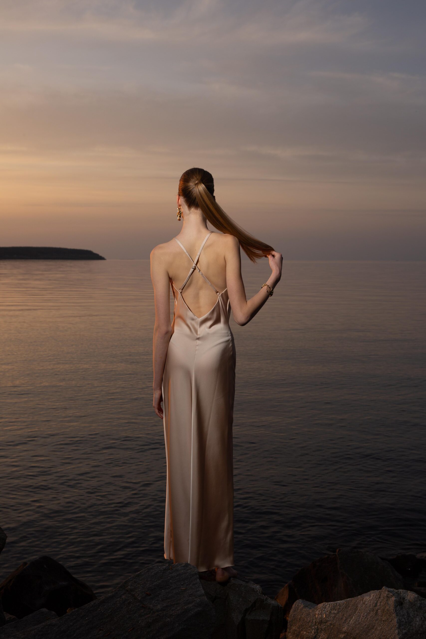 Дівчина у платті шампань кольору SLIP DRESS “LANA” від бренду Her Name стоїть спиною на фоні Чорного моря тримаючи волосся правою рукою