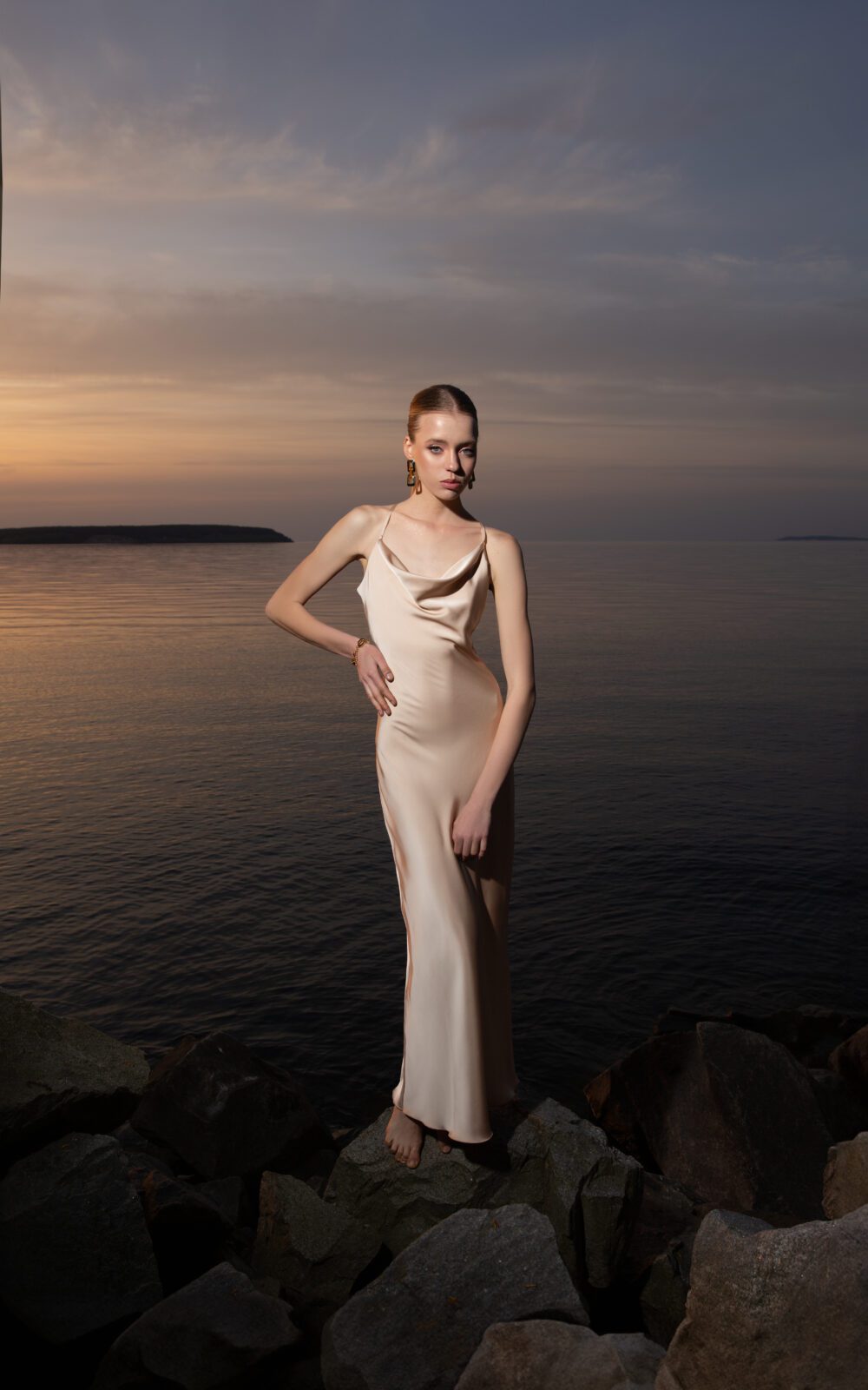 Дівчина у платті шампань кольору SLIP DRESS “LANA” від бренду Her Name стоїть на фоні Чорного моря на каміннях