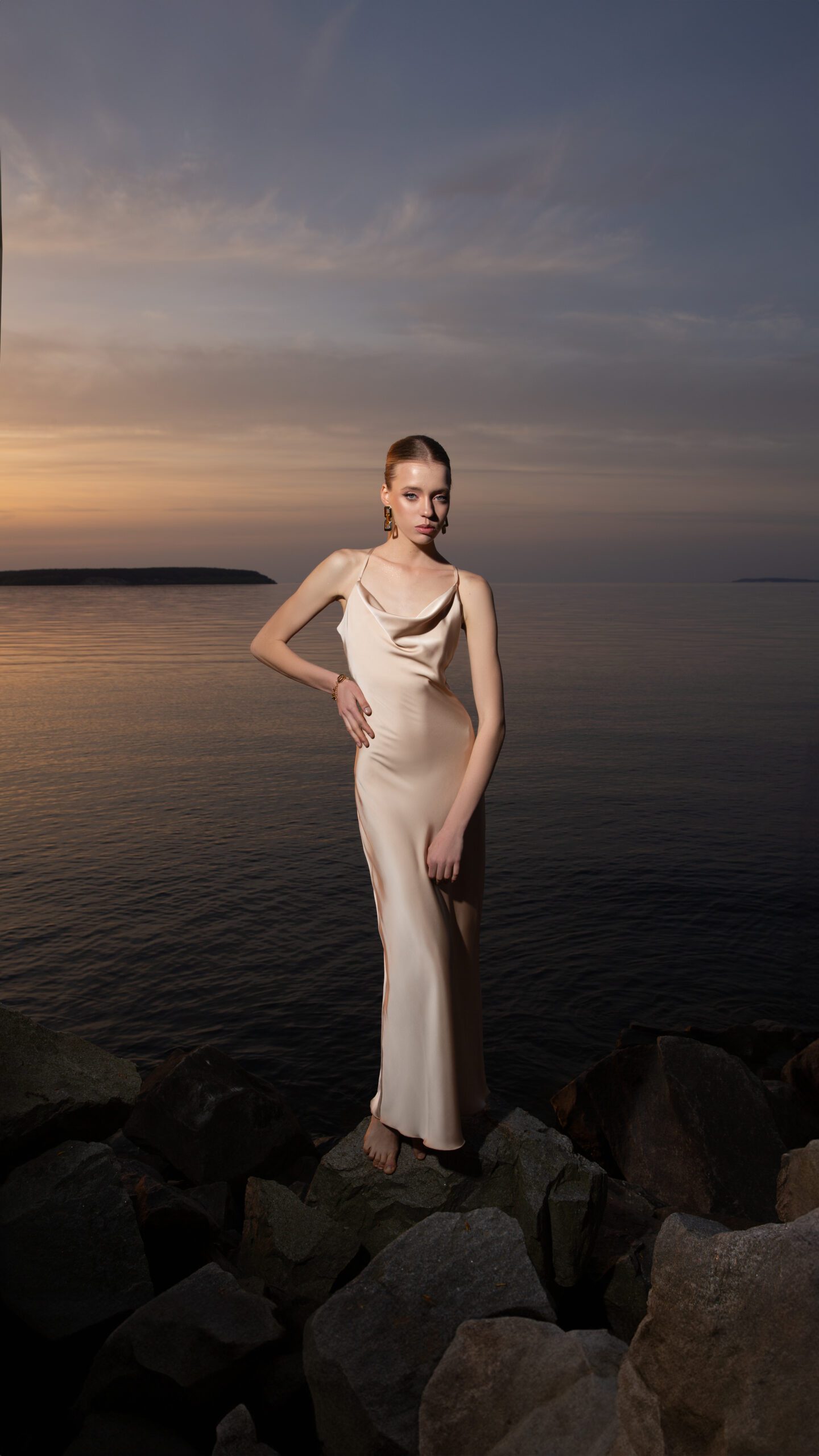 Дівчина у платті шампань кольору SLIP DRESS “LANA” від бренду Her Name стоїть на фоні Чорного моря на каміннях
