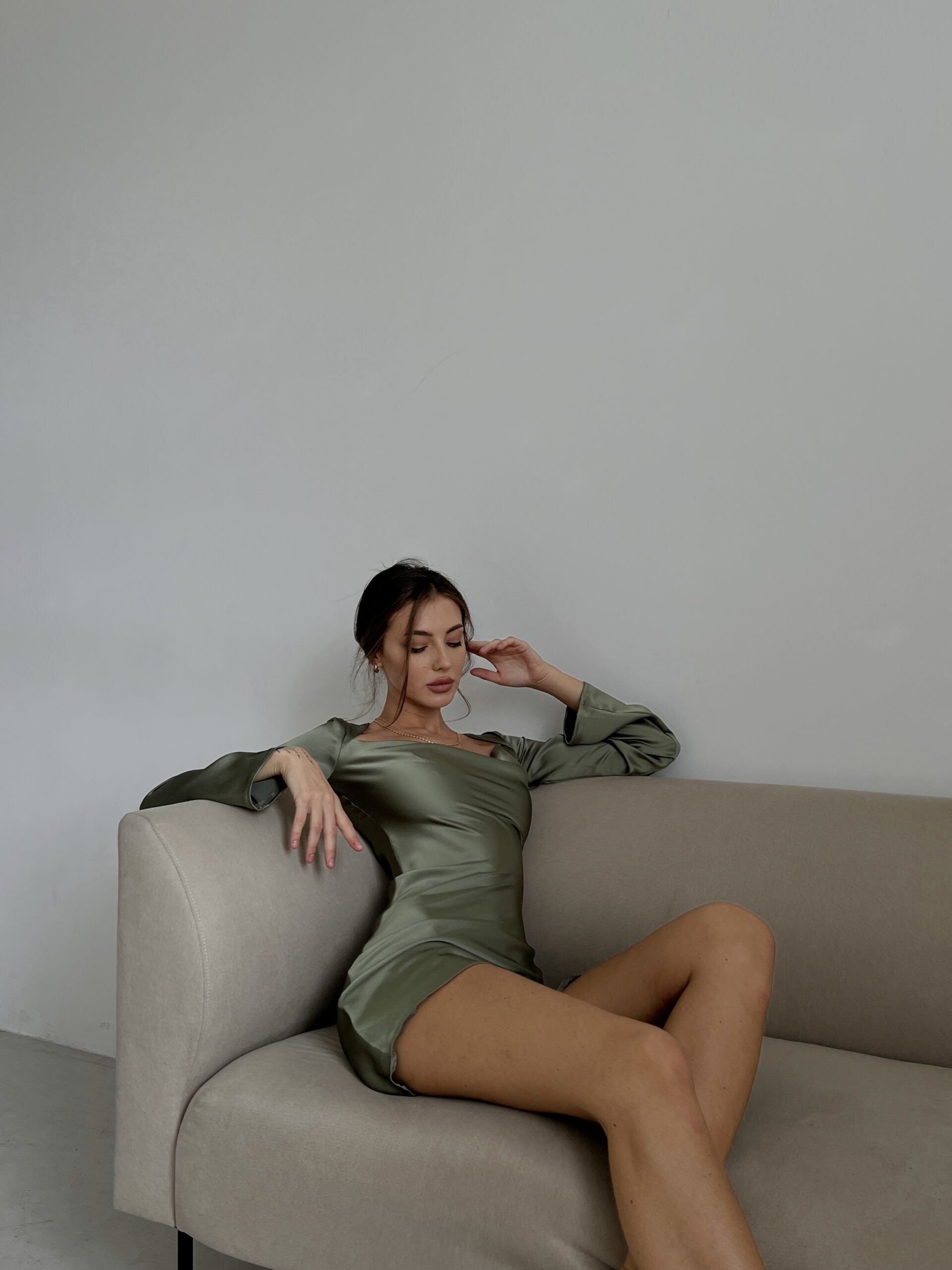 Модель у платті оливкового кольору MINI SATIN DRESS від українського бренду HER NAME сидить на дивані.