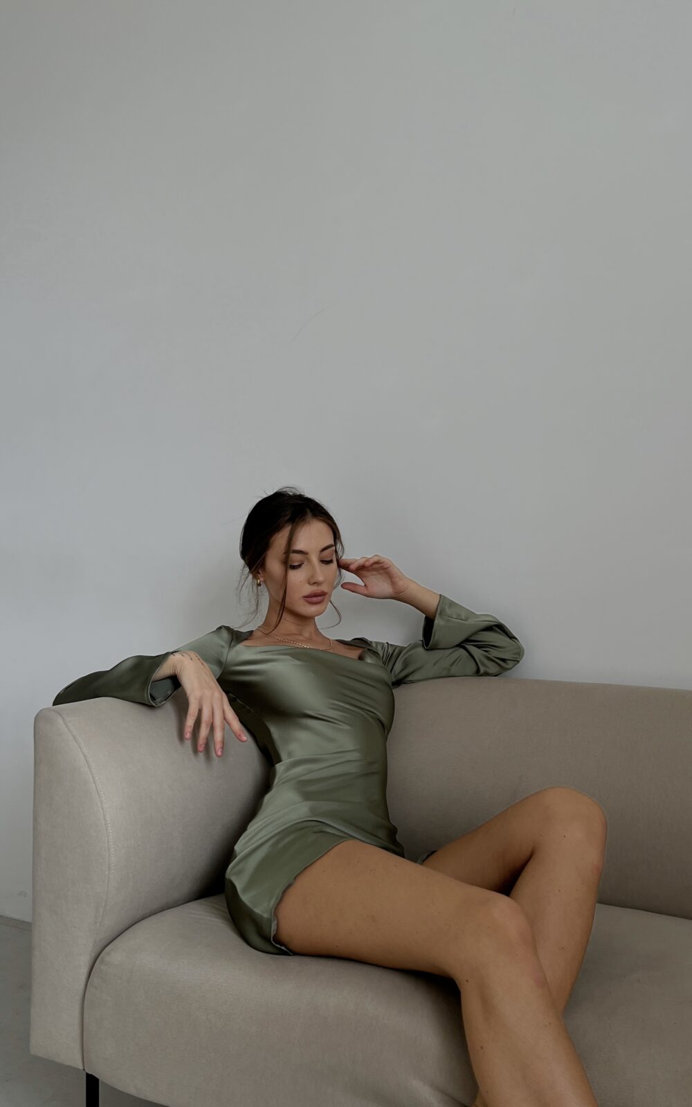 Модель у платті оливкового кольору MINI SATIN DRESS від українського бренду HER NAME сидить на дивані.